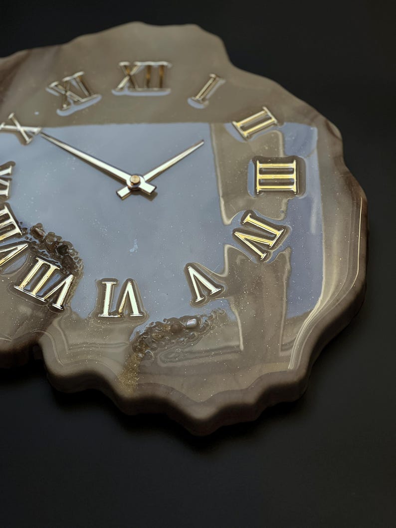 May include: A unique wall clock with a natural, organic shape. The clock face is a muted gray with gold Roman numerals and hands. The clock's edge has a marbled, brown and gold resin finish, creating a striking contrast.
