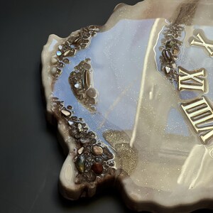 May include: A decorative resin clock with a unique, organic shape. The clock face features gold Roman numerals and is accented with embedded stones and a shimmering, pearlescent finish. The edges are a dark brown.