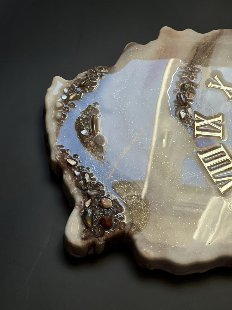 May include: A decorative resin clock with a unique, organic shape. The clock face features gold Roman numerals and is accented with embedded stones and a shimmering, pearlescent finish. The edges are a dark brown.