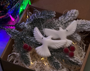 May include: Two white ceramic dove ornaments with silver hanging strings rest in a cardboard box with faux evergreen branches, red berries, and string lights. The box is filled with white paper shreds, creating a festive holiday display.