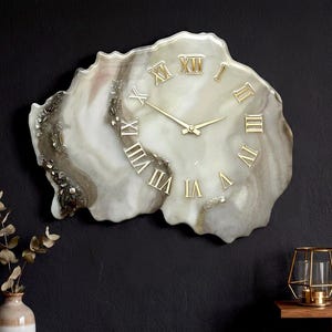 Può includere: Un orologio da parete unico con una forma marmorizzata e organica. Il quadrante dell'orologio è una miscela di bianco, grigio e marrone, con numeri romani e lancette dorate. L'orologio è montato su una parete scura, aggiungendo un tocco di eleganza.