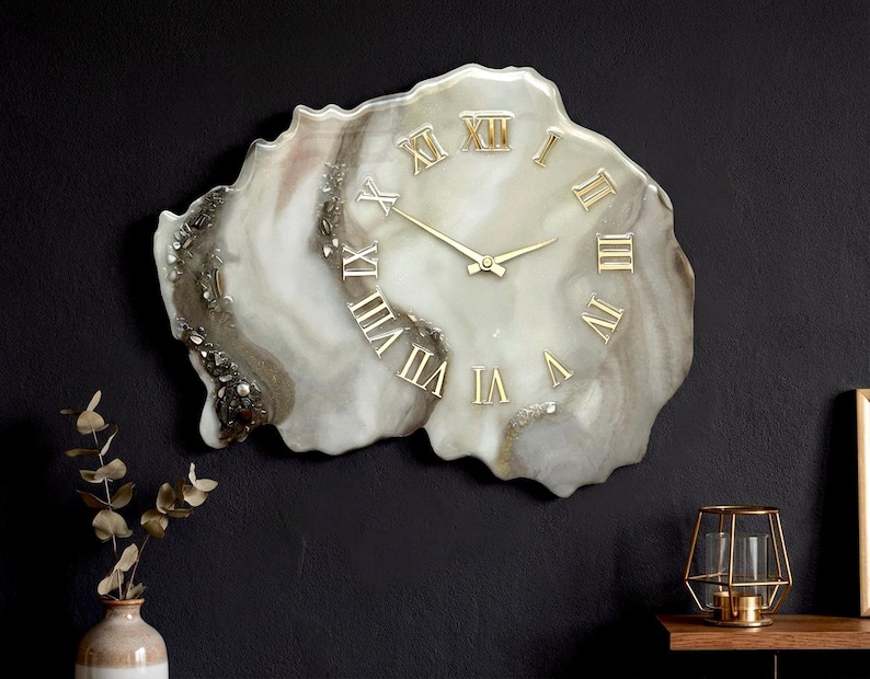 May include: A unique wall clock with a marbled, organic shape. The clock face is a blend of white, gray, and brown tones, with gold Roman numerals and clock hands. The clock is mounted on a dark wall, adding a touch of elegance to any room.