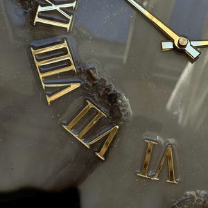 May include: Close-up of a clock face with gold Roman numerals and gold hands. The clock face is a dark gray color with a textured, possibly resin, surface. The hands point to approximately 2:15.
