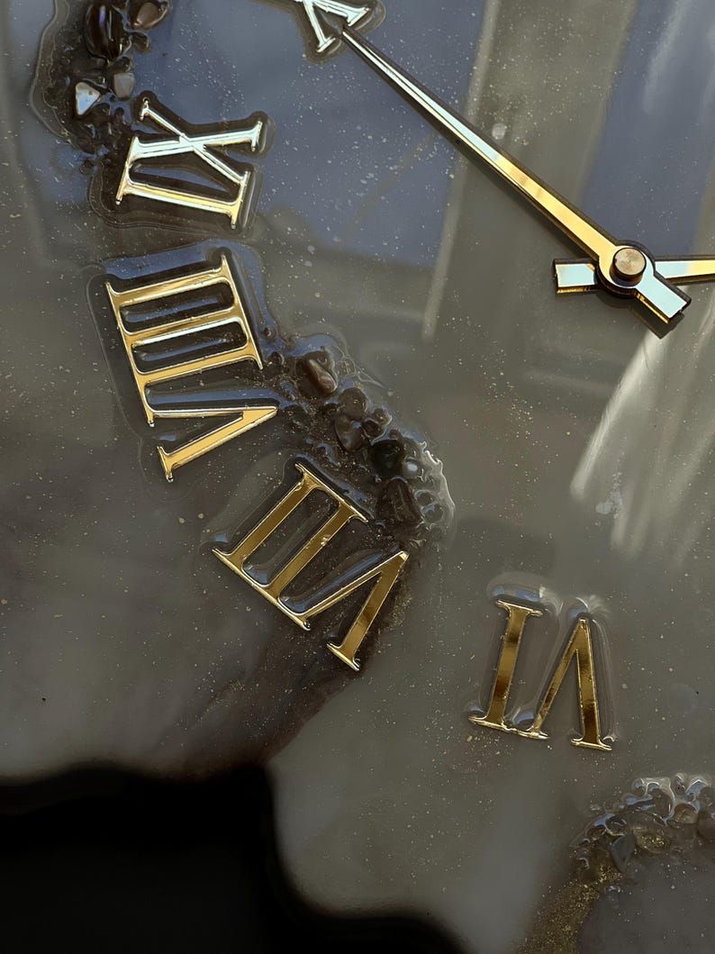 May include: Close-up of a clock face with gold Roman numerals and gold hands. The clock face is a dark gray color with a textured, possibly resin, surface. The hands point to approximately 2:15.
