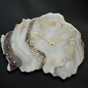 May include: A decorative wall clock with a unique, organic shape. The clock face features gold Roman numerals and hands against a marbled white and brown background. The clock is approximately 12 inches in diameter.