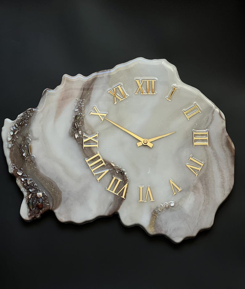 May include: A decorative wall clock with a unique, organic shape. The clock face features gold Roman numerals and hands against a marbled white and brown background. The clock is approximately 12 inches in diameter.
