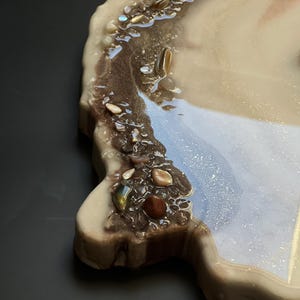May include: A decorative resin tray with a unique, organic shape. The tray features a blend of beige, brown, and shimmering blue hues, accented with embedded stones and a glossy finish. The tray is designed for decorative purposes.