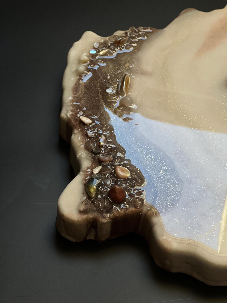 May include: A decorative resin tray with a unique, organic shape. The tray features a blend of beige, brown, and shimmering blue hues, accented with embedded stones and a glossy finish. The tray is designed for decorative purposes.