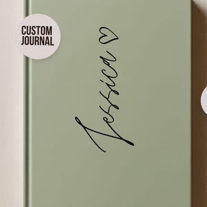 May include: Sage green custom journal with the name "Jessica" in black script. A heart symbol is above the name. The journal has a hardcover and the text "Custom Journal" is in a circle on the top left.