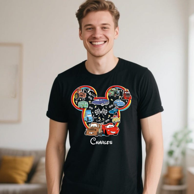 Puede incluir: Camiseta negra con un gr&aacute;fico colorido de coches de dibujos animados dispuestos en forma de cabeza de Mickey Mouse, con un arco&iacute;ris y banderas a cuadros. El nombre "Charles" est&aacute; impreso debajo.