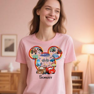 Puede incluir: Camiseta rosa claro con un dise&ntilde;o de coches de dibujos animados en forma de cabeza de Mickey Mouse. El dise&ntilde;o incluye varios veh&iacute;culos, banderas a cuadros y el nombre "Scarlett" en negro.