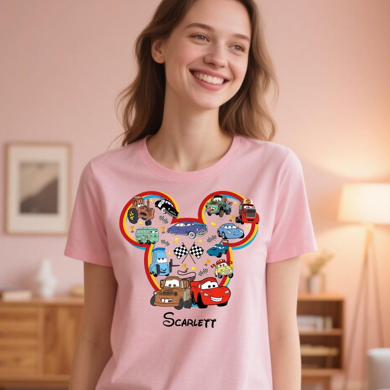 Puede incluir: Camiseta rosa claro con un dise&ntilde;o de coches de dibujos animados en forma de cabeza de Mickey Mouse. El dise&ntilde;o incluye varios veh&iacute;culos, banderas a cuadros y el nombre "Scarlett" en negro.