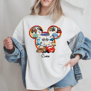 Puede incluir: Sudadera blanca con un dise&ntilde;o de dibujos animados de coches con forma de cabeza de rat&oacute;n. El dise&ntilde;o incluye varios personajes de coches, una bandera a cuadros y el nombre "Clara" en escritura negra. La sudadera tiene cuello redondo.