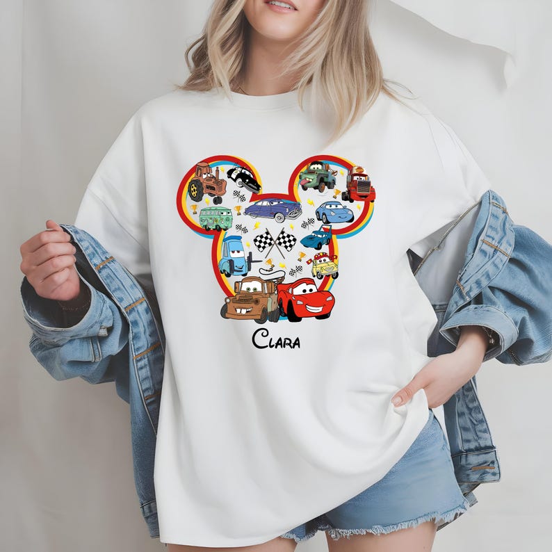 Puede incluir: Sudadera blanca con un dise&ntilde;o de dibujos animados de coches con forma de cabeza de rat&oacute;n. El dise&ntilde;o incluye varios personajes de coches, una bandera a cuadros y el nombre "Clara" en escritura negra. La sudadera tiene cuello redondo.
