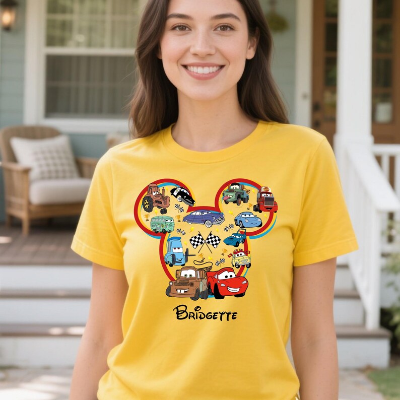 Puede incluir: Camiseta amarilla con un dise&ntilde;o de dibujos animados de coches en forma de cabeza de Mickey Mouse. El dise&ntilde;o incluye banderas de carreras y el nombre "Bridgette".