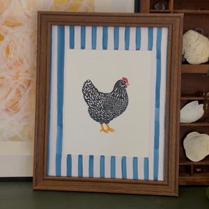 May include: Framed print featuring a black and white chicken illustration with orange feet and a red comb. The artwork is set against a white background with blue and white striped borders, encased in a brown wooden frame.