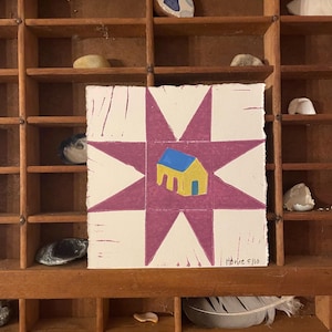 Op de afbeelding: Een vierkante kunstdruk met een paars sterrenburst-ontwerp en een geel huis met een blauw dak. Het kunstwerk is gesigneerd "Howie 5/10". De print wordt tentoongesteld in een houten organizer met diverse kleine objecten.