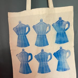May include: Cream-colored tote bag featuring a blue print of six coffee makers. The coffee maker design is repeated across the bag's surface. The bag has two cream-colored handles.