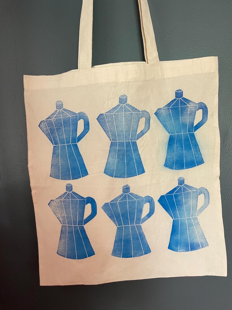 May include: Cream-colored tote bag featuring a blue print of six coffee makers. The coffee maker design is repeated across the bag's surface. The bag has two cream-colored handles.