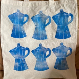May include: A white tote bag with six blue coffee pot designs. The tote bag has two handles and is made of a sturdy fabric. The coffee pot design is a geometric illustration of a stovetop espresso maker.