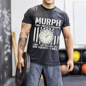May include: A black short-sleeve t-shirt featuring a white graphic of a stopwatch centered over a distressed American flag. The text reads MURPH, 1 MILE, 100 PULLUPS, 300 SQUATS, and 1 MILE. The design is printed in a weathered, off-white font style.