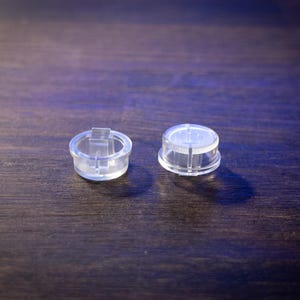 May include: Two transparent plastic caps are shown on a dark wooden surface. One cap has a rectangular tab, the other is a simple, rounded design. The caps are clear, allowing a view of the surface beneath.