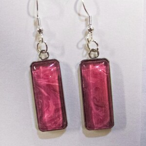May include: A pair of dangle earrings featuring rectangular, magenta resin pendants with a marbled design. The pendants are framed in a dark metal and suspended from silver-coloured fish hook ear wires.