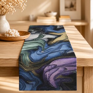 May include: A rectangular table runner featuring a fluid, marbled pattern in shades of deep blue, purple, sage green, and gold. The fabric displays swirling, abstract liquid-like textures that span the entire length of the textile surface.