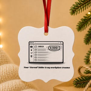 May include: A white, scalloped-edge ornament hanging from a red ribbon. The center features a black graphic of an email inbox showing 9,999+ unread messages. Below the graphic, the text reads, Your 'Unread' folder is my workplace trauma.