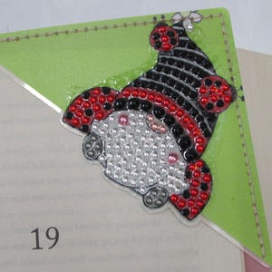 May include: A handmade corner bookmark featuring a ladybug design. The bookmark has a green background with a ladybug character made of red, black, and silver rhinestones. The bookmark is placed on the corner of a book.