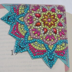 May include: A colorful, beaded corner bookmark featuring a mandala design. The bookmark has a turquoise border and is adorned with pink, yellow, and orange beads. It is placed on the corner of a book, with the page number 19 visible.