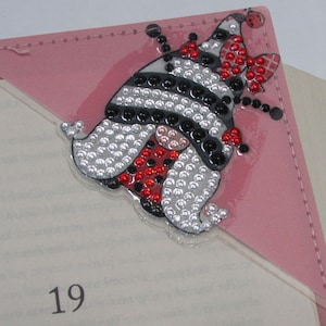May include: A pink leather bookmark with a decorative gnome design. The gnome is adorned with silver, black, and red rhinestones. The bookmark is placed on a book, with the number 19 visible.