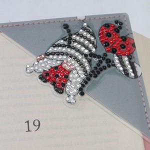 May include: A decorative corner bookmark featuring a butterfly and ladybug design. The butterfly has black and white stripes with red and silver accents. The ladybug is red with black spots. The bookmark is gray with pink trim and is placed in a book.