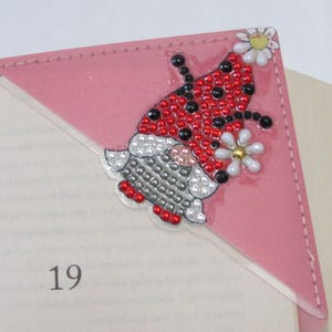 May include: Pink corner bookmark with a ladybug design. The ladybug has red and black spots, with a white and gold flower accent. The bookmark is made of pink material and is placed on a book.