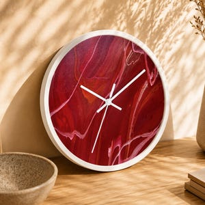 Puede incluir: Un reloj de pared redondo con marco blanco y manecillas blancas. La esfera presenta un diseño de arte fluido abstracto con patrones de remolinos en rojo intenso, burdeos y finas líneas blancas. El acabado es brillante y no tiene números.