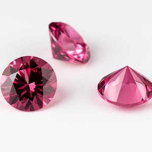 May include: Three faceted, pink gemstones of varying shapes are displayed against a white background. The gemstones have a brilliant cut, reflecting light. One is round, one is pear-shaped, and one is a triangular shape.