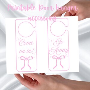 Może przedstawiać: Różowy, drukowany dodatek do wieszaka na drzwi. Projekt przedstawia dwa wieszaki na drzwi z napisami "Come on in!" i "Go Away" w kursywie. Każdy wieszak ma kokardę i dekoracyjne iskierki.