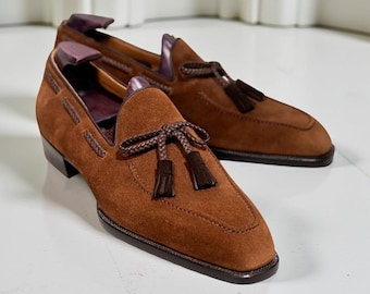 Könnte beinhalten: Ein Paar braune Wildleder-Loafer mit Quasten-Details. Die Schuhe haben eine eckige Spitze und einen niedrigen Absatz. Die Loafer werden auf einer weißen Oberfläche präsentiert.