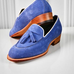 May include: A pair of blue suede loafers with a tassel detail. The shoes have a light brown sole and heel, with white stitching and trim. The shoes are stacked on top of each other.