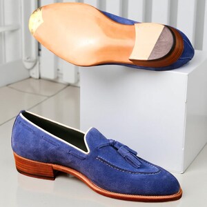 May include: Blue suede loafers with a tassel detail and a stacked heel. The shoe has a leather sole and a white trim. The second shoe is displayed upside down, showing the sole.