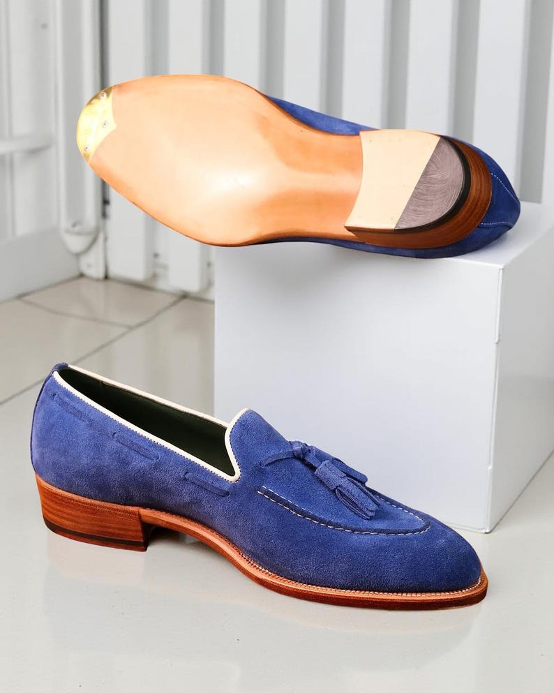 May include: Blue suede loafers with a tassel detail and a stacked heel. The shoe has a leather sole and a white trim. The second shoe is displayed upside down, showing the sole.
