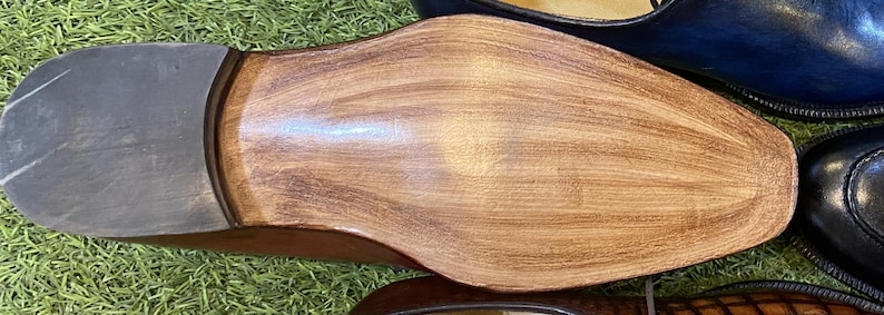 May include: Close-up of the sole of a brown leather shoe, showing the grain and texture of the wood. The heel is a darker gray color. Other shoes are visible in the background.