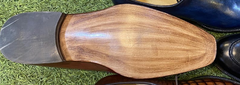 May include: Close-up of the sole of a brown leather shoe, showing the grain and texture of the wood. The heel is a darker gray color. Other shoes are visible in the background.