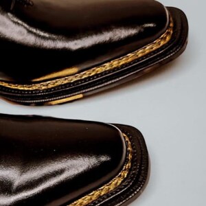 May include: Close-up of two black leather dress shoes. The shoes have a square toe design and a gold-coloured trim around the sole. The leather has a glossy finish, reflecting light. The shoes are likely for formal occasions.