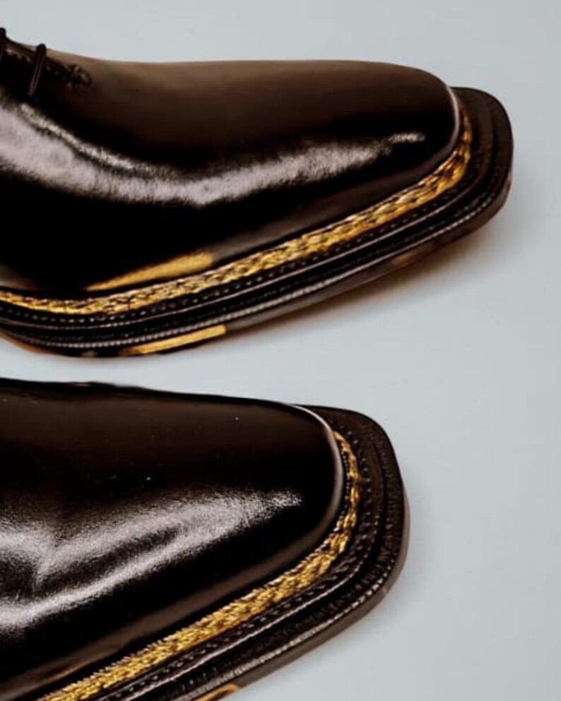 May include: Close-up of two black leather dress shoes. The shoes have a square toe design and a gold-coloured trim around the sole. The leather has a glossy finish, reflecting light. The shoes are likely for formal occasions.