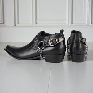 Puede incluir: Botines de cuero negro con puntera afilada, hebillas plateadas y detalles de cadena. Las botas tienen un tacón bajo y un diseño de inspiración western. Las botas se exhiben sobre una superficie gris clara.