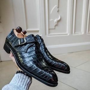 Könnte beinhalten: Ein Paar schwarze Lederschuhe mit Krokodilleder-Textur. Die Schuhe haben einen Schnallenverschluss und einen niedrigen Absatz. Die Schuhe werden vor einem hellen Hintergrund präsentiert. Die Schuhe sind ein klassischer Stil.