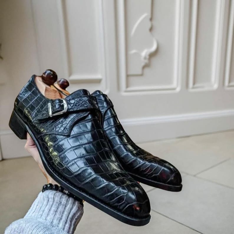 May include: A pair of black leather shoes with a crocodile skin texture. The shoes have a buckle fastening and a low heel. The shoes are presented against a light background. The shoes are a classic style.