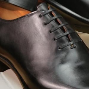 May include: Close-up of a black leather dress shoe with a classic lace-up design. The shoe has a polished finish and a subtle sheen, suggesting high-quality craftsmanship. The laces are black, and the shoe appears to be new.