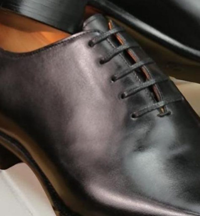 May include: Close-up of a black leather dress shoe with a classic lace-up design. The shoe has a polished finish and a subtle sheen, suggesting high-quality craftsmanship. The laces are black, and the shoe appears to be new.